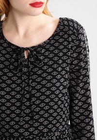 Vestido negro con un patrón de diamantes en blanco, con un escote redondo y un detalle de lazo en forma de ojal en la parte delantera. Mangas largas con cintura ajustada.