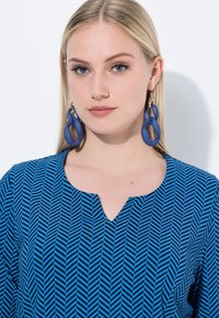 Blusa azul con patrón, de escote en V y diseño de espiga, combinada con grandes pendientes de aro azules mate. Textura de tela suave.