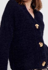 Navy blue, oversized cardigan made of fuzzy material with a v-neck and four prominent gold-colored buttons down the front.