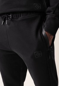 Zwarte joggings met een trekkoord in de taille, voorzien van ritszakken aan de zijkant en kleine logo-accenten. Textuurstof met een gladde afwerking.