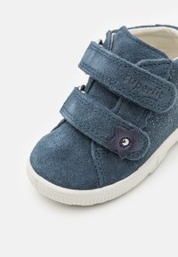 Tmavě modrá semišová bota se dvěma suchými zipy, hvězdicový akcent s knoflíkem, bílá gumová podrážka a strukturovaný povrch.