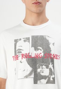 Nærbillede af en hvid T-shirt med sort-hvide portrætter og rød tekst, der står "THE ROLLING STONES" på tværs af brystet.