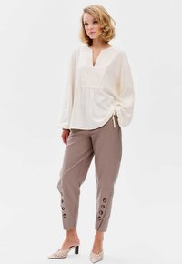 Weiße Bluse mit V-Ausschnitt und elastischen Bündchen, kombiniert mit taubfarbenen Hosen, die Knopfakzente an den Knöcheln aufweisen, und offenen Schuhen.