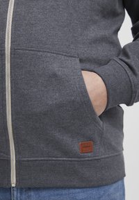 Felpa grigia con zip realizzata in tessuto morbido. Presenta tasche frontali, polsini a costine e una piccola etichetta in pelle marrone sul bordo inferiore.