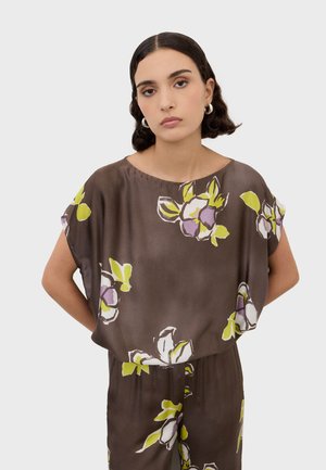 Jeune femme aux cheveux courts et foncés portant un chemisier marron à motif floral et un pantalon assorti, debout avec les mains derrière le dos sur un fond neutre.