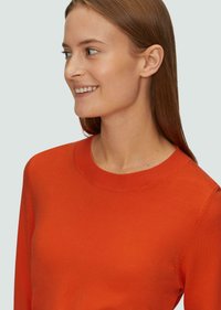 Czerwony sweter z dzianiny o okrągłym dekolcie, z długimi rękawami i gładką teksturą, noszony z rozpuszczonymi włosami i subtelnym uśmiechem.