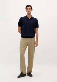 Marine polo t-shirt, kortærmet, to-knaps placket; parret med lyse beige linnedbukser; sorte snørebåndssko. Ensfarvet baggrund.