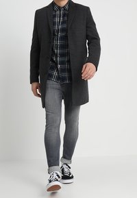 Cappotto di lana grigio scuro con una vestibilità su misura, indossato sopra una camicia a quadri blu navy, abbinato a jeans slim grigi chiari e sneakers nere.