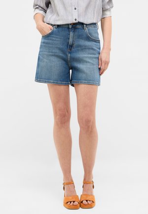 Shorts de mezclilla en un lavados azul medio con cintura alta, que cuentan con cinco bolsillos y dobladillos rematados. Combinados con sandalias gruesas de tacón naranja.
