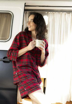 Frau in roter karierten Pyjamas, die eine Tasse hält und sich an eine Wohnwagentür mit weißen Vorhängen im natürlichen Licht lehnt.