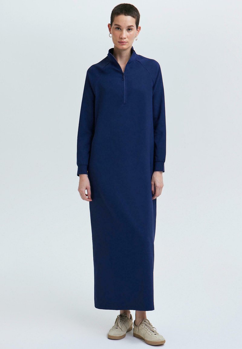 Personne portant une longue robe bleu marine à manches longues, avec un col à moitié zippé, et des baskets beiges, se tenant devant un fond clair.