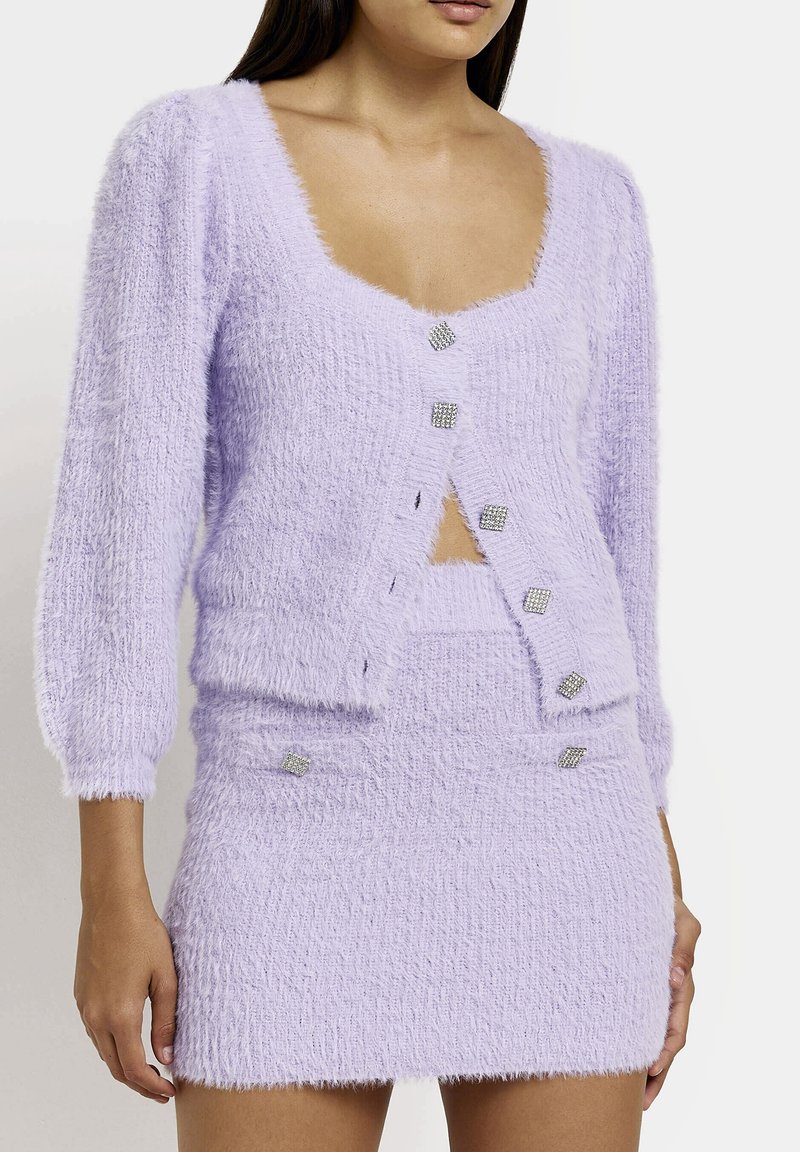 Gilet en lilas duveteux avec des boutons en strass carrés, associé à une mini-jupe assortie avec une texture et des accents de boutons similaires.