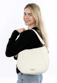 White leather shoulder bag with a curved shape, featuring a prominent logo and adjustable strap, held by a person in a black sweater.