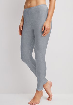 Vrouw draagt nauwsluitende lichtgrijze legging, staat blootsvoets op een witte vloer tegen een lichtgrijze achtergrond.