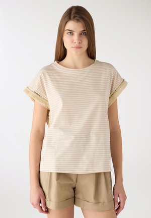 Giovane donna con capelli castani lunghi che indossa una maglietta a maniche corte beige a righe e pantaloncini kaki, in piedi davanti a uno sfondo semplice.