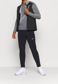 Zwarte Nike-jas met gestreepte lange mouwen, gecombineerd met een grijs gestreept shirt en zwarte broek. Witte Nike-schoenen, minimale hardware.
