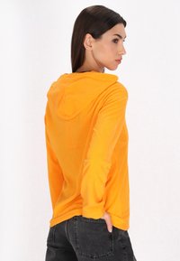 Felpa arancione con cappuccio realizzata in tessuto morbido, caratterizzata da maniche lunghe e una vestibilità comoda. Cuciture visibili sulle maniche e sull'orlo.