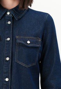Chemise en denim avec fermeture à boutons sur le devant et poche à rabat avec bouton, portée par une personne ayant les cheveux noirs courts.
