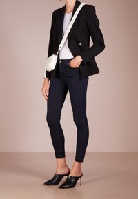 Persoon in een zwarte blazer, witte shirt, donkere skinny jeans, zwarte hoge muilriemen en witte crossbody tas, staande tegen een effen achtergrond.