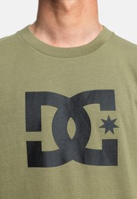 Camiseta de algodón verde oliva con un gran logo negro de DC con una estrella. Diseño de cuello redondo, textura suave y mangas cortas.