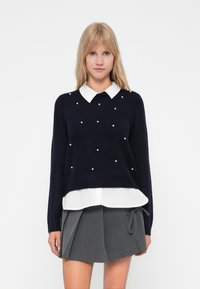 Marineblauwe trui met witte kraag, met parelachtige accenten op de voorkant. Gelaagd over een wit shirt en gecombineerd met een grijze plooienrok.