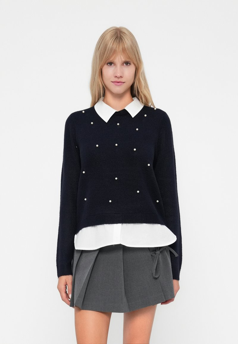 Marineblauwe trui met witte kraag, met parelachtige accenten op de voorkant. Gelaagd over een wit shirt en gecombineerd met een grijze plooienrok.