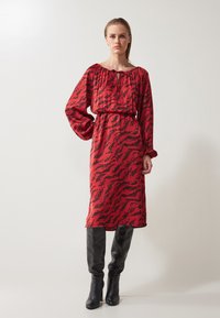 Red dress with an abstract black pattern, long sleeves, gathered neckline, and a cinched waist, paired with knee-high black boots.