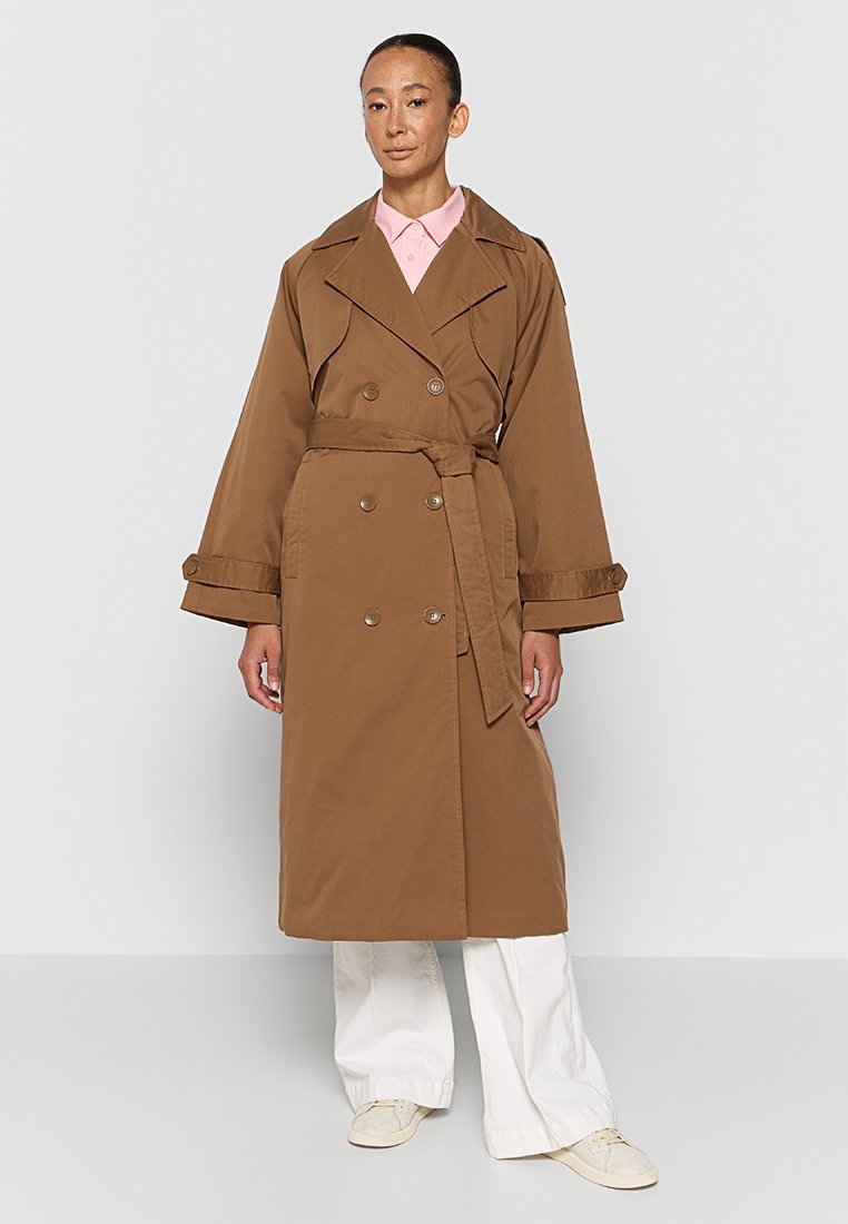Manteau trench marron à double boutonnage avec une ceinture à nouer, grands revers et détails de boutons, porté sur une chemise rose à col et un pantalon blanc.