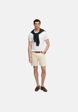 Man draagt wit T-shirt, beige korte broek, bruine riem en schoenen, met zwarte trui over de schouders gedrapeerd, staand tegen een witte achtergrond.