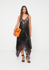 Vestido maxi preto com padrão e detalhes florais, com bainha assimétrica e alças finas. Combinado com uma clutch laranja e saltos pretos.