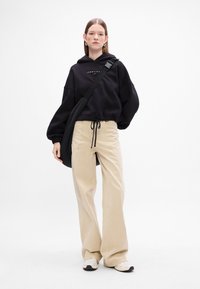 Felpa nera con cappuccio e logo, pantaloni a gamba larga beige e borsa a tracolla nera. Il modello indossa sneakers bianche con dettagli neri.