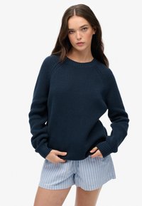 Ciemnoniebieski sweter o teksturowym wykończeniu z prążkowanymi mankietami i dołem, w zestawieniu z jasnoniebieskimi spodniami w białe paski. Krój jest luźny i swobodny.