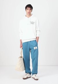 Balta gobtuvė, aprašyta Calvin Klein logotipu, derinama su mėlynomis kelnėmis, turinčiomis baltą etiketę, ir kreminės spalvos rankine. Rudi ir balti sportiniai batai.