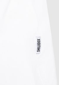 Tissu en coton blanc avec une texture lisse. Présente une étiquette noire cousue avec le texte "SMILODOX" sur le côté gauche. Design minimaliste.