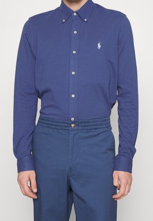 Homme portant une chemise bleue ajustée à manches longues avec un petit logo blanc brodé sur la poitrine et un pantalon bleu assorti.