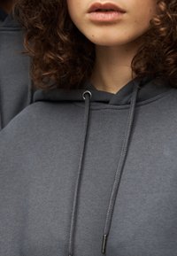 Γκρι βαμβακερό φούτερ με κορδόνι και μεταλλικά τελειώματα, άνετη εφαρμογή και απαλή υφή. Κοντινό πλάνο στη λαιμόκοψη και το πρόσωπο.