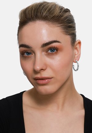 Silberne Creolen, glatte Textur, kreisförmiges Design; kombiniert mit einem schwarzen Rippenshirt. Klare blaue Augen und natürliche Schminke.