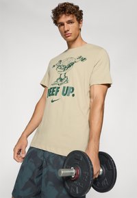Beige t-shirt med grøn grafisk design af en hamburger og teksten "BEEF UP." Sort vægtstang i den ene hånd, parret med mønstrede shorts.
