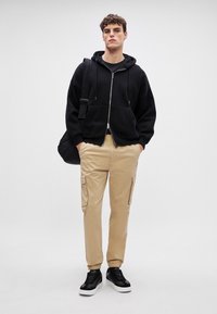 Zwarte zip-up hoodie met voorzakken, beige cargobroek met zijzakken en zwarte sneakers met witte zolen.