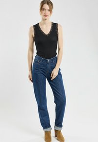 Haut sans manches noir avec bord en dentelle, associé à un jean en denim bleu taille haute. Le mannequin porte des bottines marron ; fond uni.