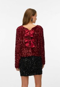 Rød palietbluse med lange ærmer og to store satinbuer bagpå. Kombineret med en sort palietnederdel. Glat tekstur.