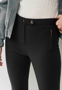 Černé přiléhavé kalhoty s hladkým povrchem. Obsahují dvě dekorativní knoflíky na pase a kapsu na zip po straně.