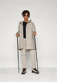 Ανοιχτό μπεζ αδιάβροχο μπουφάν με κουκούλα, συνδυασμένο με ταιριαστά παντελόνια, μαύρο πουκάμισο, γυαλιά ηλίου και μπαστούνια πεζοπορίας. Υφασμάτινη υφή, σύγχρονος σχεδιασμός.