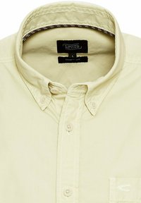 Jasno beżowa koszula z guzikami, klasyczny kołnierzyk, dwa guziki z przodu, teksturowany materiał oraz małe logo na kieszeni piersiowej.