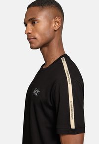 Man i svart EA7 Emporio Armani t-shirt med beige varumärkesrand på axeln, vänd mot vänster mot en enkel ljus bakgrund.