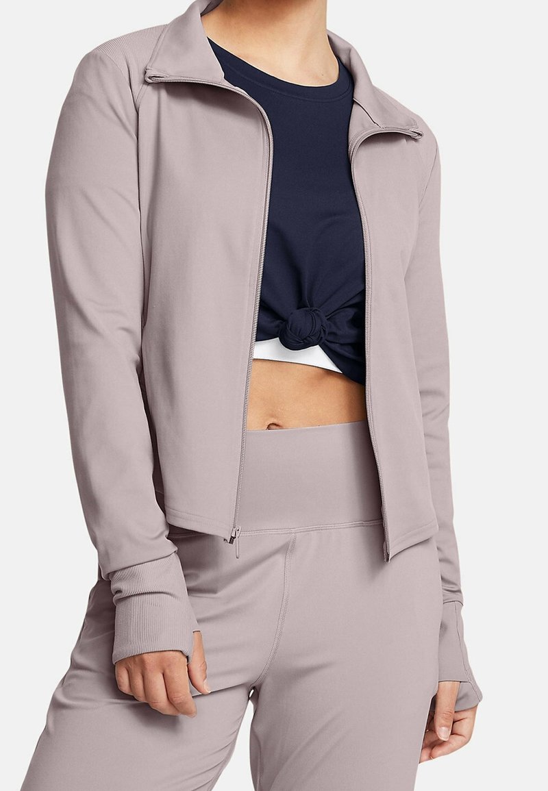 Femme portant une veste zippée mauve clair et des leggings assortis sur un crop top noir noué laissant apparaître le ventre.