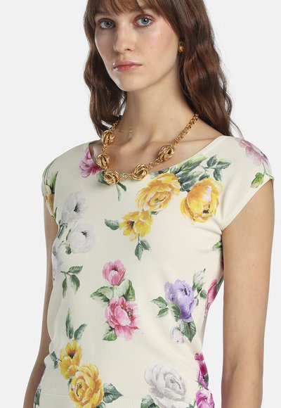 Frau trägt ein kurzärmeliges Oberteil mit gelben, pinken, violetten und weißen Blumen sowie eine klobige goldene Halskette vor weißem Hintergrund.