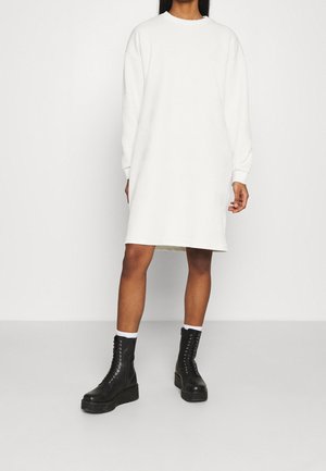 Kobieta ubrana w długą białą, prążkowaną, sweterkową sukienkę oraz czarne, sznurowane botki na platformie, stojąca na tle jasnego, jednolitego tła.