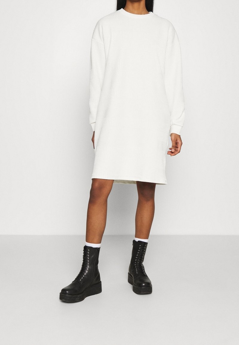 Femme portant une longue robe pull blanche côtelée et des bottines plates-formes noires à lacets, debout devant un fond clair uni.