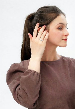 Blouse en velours côtelé marron à manches bouffantes, portée par une personne touchant son oreille. Bague en argent au doigt, fond neutre.